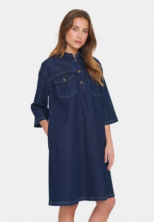 Jeansskjol i mörkblått med krage, två bröstfickor, knäppning fram och trekvartslång ärm. Slät textur.