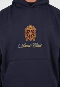 Granatowy hoodie z dużą haftowaną emblematyką w złotym i burgundowym kolorze, z napisem „Social Club” w niebieskim kursywie nad kieszenią typu kangur.