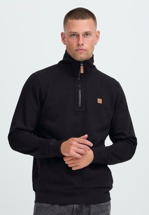Mann mit kurzen Haaren, der einen schwarzen Pullover mit Viertelreißverschluss und dunklen Hosen trägt, und vor einem schlichten hellen Hintergrund steht.
