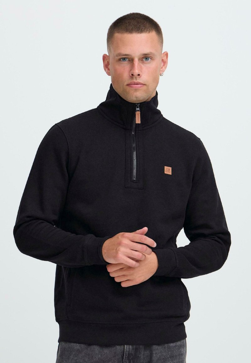 Uomo con i capelli corti che indossa un maglione a zip nera e pantaloni scuri, in piedi davanti a uno sfondo chiaro e semplice.