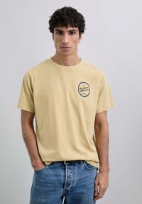 Joven con cabello rizado y oscuro, vistiendo una camiseta beige con un pequeño logo y jeans azules, de pie frente a un fondo claro y liso.