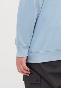 Světle modrá mikina s žebrovanými manžetami a spodním lemem, měkká textura, těsný střih, část tmavého outfitu, ukazující boční pohled na rukáv a ruku.