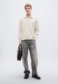 Μπεζ ριμπ πλεκτό πουλόβερ με γιακά, συνδυασμένο με φαρδιά γκρι τζιν. Το μοντέλο κρατά μαύρη δερμάτινη τσάντα. Απλά παπούτσια. Ουδέτερο φόντο.