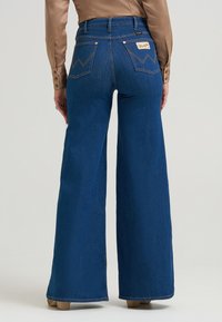 Υψηλής μέσης μπλε τζιν παντελόνι σε καμπάνα με λεία υφή, εμφανή ραφές και δύο πίσω τσέπες με σχέδιο ζιγκ-ζαγκ.