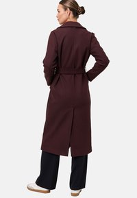 Vrouw met een bun, gekleed in een donkerburgundy jasje met ceintuur, zwarte broek en witte sneakers, staand met haar handen op haar heupen, zijwaarts kijkend.