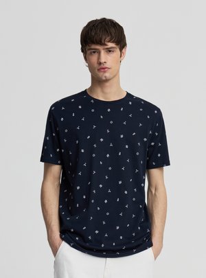 Giovane uomo con indosso una maglietta blu navy con piccoli simboli nautici bianchi, in piedi con le mani nelle tasche su uno sfondo grigio uniforme.