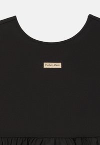 Musta kleit, mis on valmistatud sujuvast ja venitavast kangast, millel on ümar kaelus. Esiküljel, volangiga krae kohal, asub Calvin Kleini logo silt.