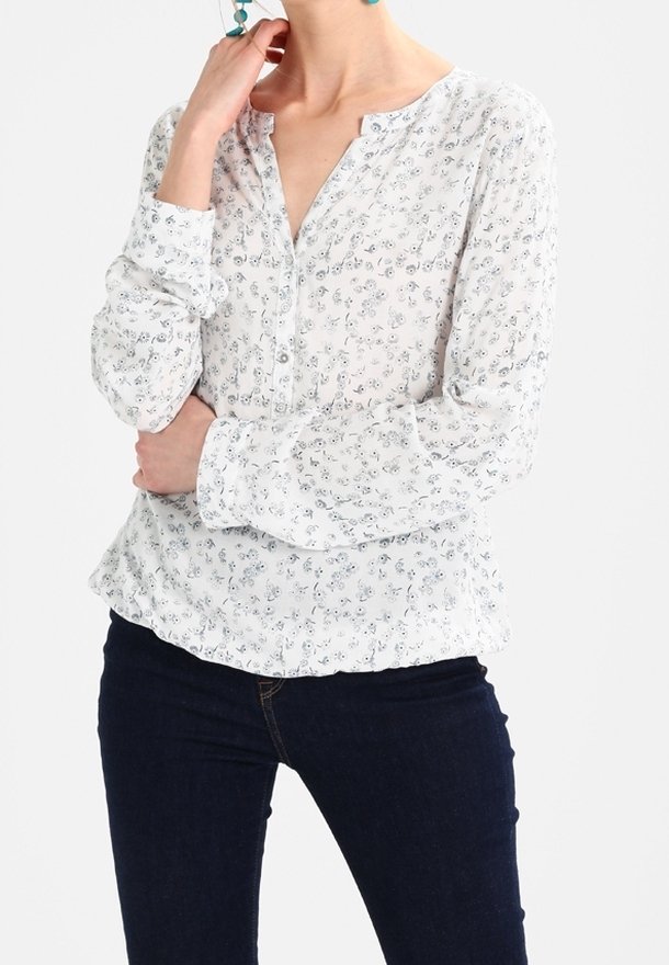 Blouse blanche à manches longues avec imprimé floral, dotée d'un col cranté et d'une fermeture par boutons. Tissu léger avec une légère texture, associée à un jean foncé.