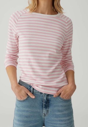 Personne portant une chemise à manches longues rayée rose et blanche rentrée dans un jean en denim bleu clair, les mains dans les poches.