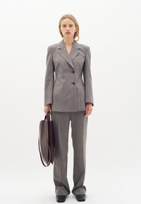 Grå skräddarsydd kostym med vida byxor, texturerad tyg och en strukturerad kavaj med notch-lapel och två knappar. Håller en brun väska.