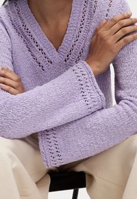Maglione lavorato a maglia di un leggero color lavanda con scollo a V, caratterizzato da motivi a punto aperto e maniche larghe, abbinato a pantaloni beige.