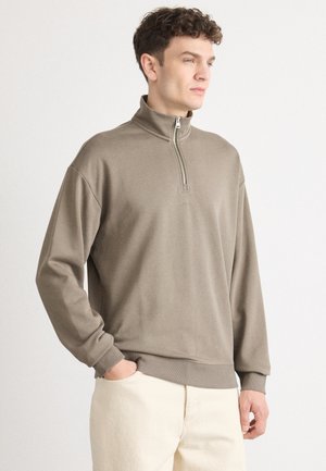 Sweatshirt marron clair avec un col à demi-zip, des poignets et un ourlet côtelés. Présente un branding subtil sur la poitrine et une coupe décontractée.