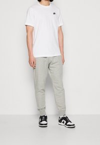 Hombre vestindo una camiseta blanca de Nike, pantalones de chándal gris claro y zapatillas Nike en blanco y negro, de pie contra un fondo blanco liso.
