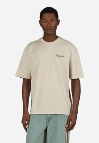 Beżowa bawełniana koszulka z krótkim rękawem, okrągłym dekoltem i czarnym haftowanym logo "Pegador" na lewej piersi. Luźny krój.
