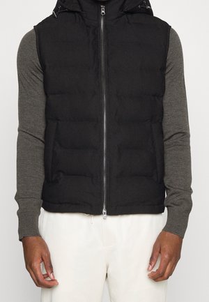 Un mannequin porte un gilet matelassé noir à capuche par-dessus un haut gris à manches longues et un pantalon blanc. Le gilet est doté d’une fermeture éclair sur le devant et de poches latérales.