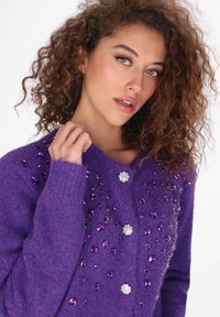 Lila kardigán, texturált anyagból készült, különböző lila árnyalatú és ezüst részletekkel díszítve, mint drágakövek. Elöl gombos záródás.