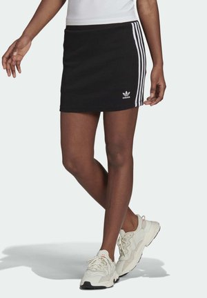 Osoba nosząca czarną mini spódniczkę Adidas z białymi paskami i białe trampki, stojąca ze skrzyżowanymi nogami na jasnym tle.