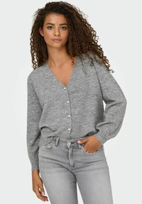 Cardigan gris en tissu tricoté doux avec un col en V, un front à boutons avec des boutons ressemblant à des perles, et des manches larges et effilées. Porté avec un jean gris clair.