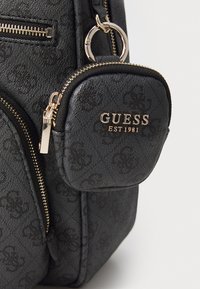 Zaino nero con texture, dotato di hardware dorato, un piccolo astuccio con zip e un motivo in rilievo. L'etichetta del marchio recita "GUESS EST 1981."