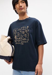 Σακάκι μπλε ναυτικού βαμβακερή μπλούζα με χρυσό σχέδιο γραφικών που περιλαμβάνει διάφορα σύμβολα και κείμενο. Έχει κοντά μανίκια και άνετη εφαρμογή, συνδυασμένο με μια ελαφριά τσάντα.