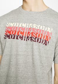 Grå kortærmet t-shirt med et flerfarvet tekstdesign, hvor "SCOTCH & SODA" står skrevet i forskellige fede skrifttyper og størrelser henover brystet.