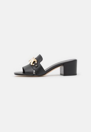 Mule noire à bout ouvert avec talon bloc moyen et grande boucle décorative dorée sur une large lanière en cuir.