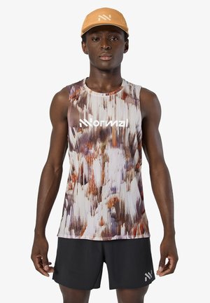 Junger Mann mit gemustertem ärmellosem Hemd, schwarzer Shorts und beiger Kappe, stehend vor hellem einfarbigem Hintergrund.