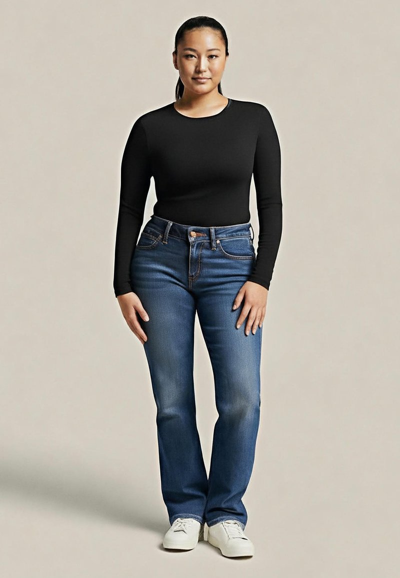 Donna in piedi con una maglietta nera aderente a maniche lunghe, jeans blu e sneakers bianche contro uno sfondo beige semplice.