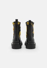 Botas de cuero negro hasta el tobillo con suelas gruesas y paneles de tela en un patrón barroco amarillo ornamental, vistas desde atrás sobre un fondo blanco.