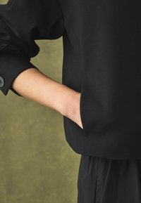 Must mustreiga lõdva lõikega, küljetasku ja nööpidega mansetiga. Sile tekstuur ja minimalistlik disain.