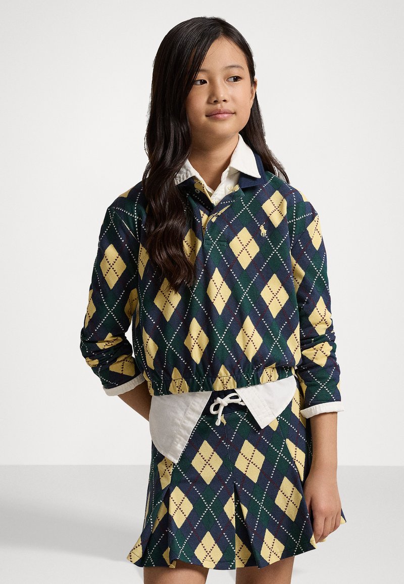 Többszínű, navy, zöld és sárga argyle mintás pulóver, fehér galléros inggel és harmonizáló, pliszírozott szoknyával párosítva.