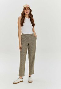 Kobieta z długimi falującymi włosami, nosząca beżową czapkę, bezrękawnik biały top, kraciastą prostą spódnicę i białe sneakersy, pozująca z rękami w kieszeniach.