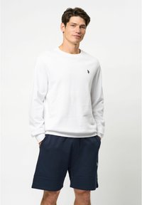 Witte lange mouwen t-shirt met een ronde halslijn, met een klein zwart logo op de borst. Gecombineerd met een marineblauwe korte broek.