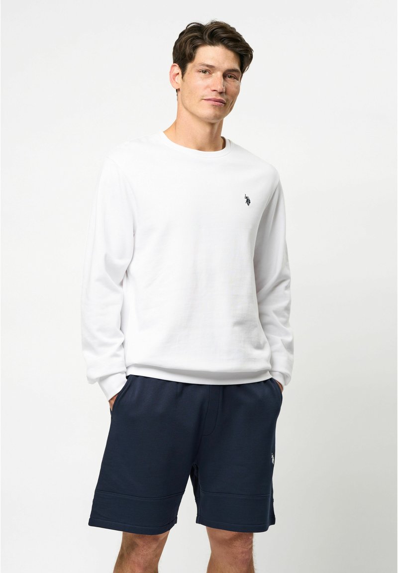 Witte lange mouwen t-shirt met een ronde halslijn, met een klein zwart logo op de borst. Gecombineerd met een marineblauwe korte broek.