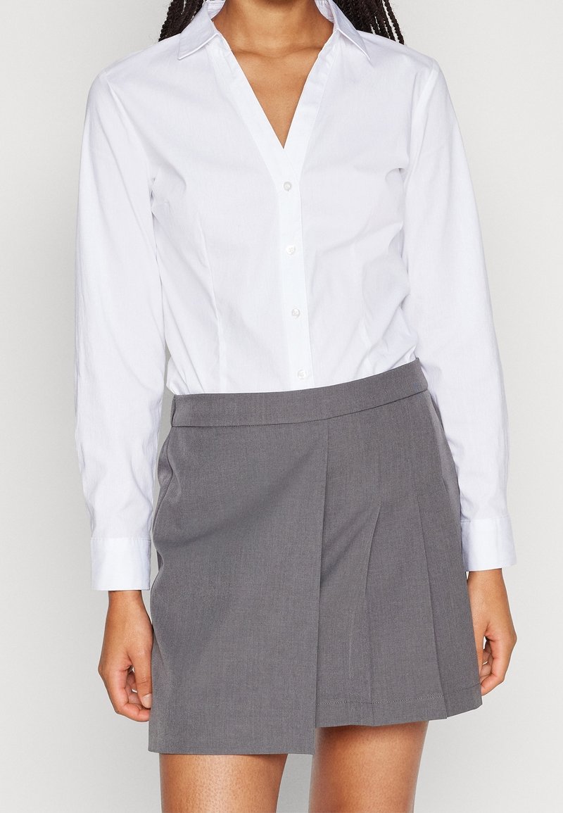 Femme portant une chemise blanche à manches longues boutonnée, rentrée dans une jupe plissée grise taille haute, debout devant un fond uni.