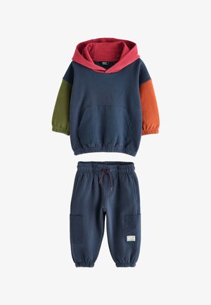 Dwuczęściowy strój dziecięcy z granatową bluzą z kapturem w czerwonym kolorze, zielonymi i pomarańczowymi rękawami oraz pasującymi granatowymi spodenkami dresowymi z naszywanymi kieszeniami.