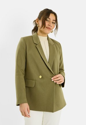 Mujer con cabello oscuro recogido en una coleta baja, lleva pendientes de aro dorados, blazer verde oliva con botón floral, camiseta crema y pantalones blancos.