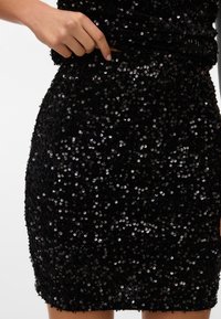 Main soulevant la taille d'une jupe noire brillante couverte de sequins portée sur la partie inférieure du corps sur fond blanc.