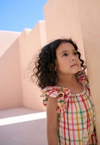 Colorful checkered dress with ruffled sleeves in pink, yellow, green, and white; child looking up against a light pink wall background.