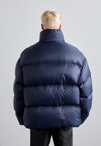 Person mit kurzem blonden Haar, der eine übergroße marineblaue Daunenjacke trägt und mit dem Rücken zu einem neutralen grauen Hintergrund steht.