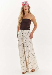 Žena stojaca v bezramienkovom tmavo hnedom topu, širokých bielych nohaviciach s čiernymi bodkami, kvetinovej šatke na hlave, náhrdelníku a sandáloch na jednoduchom pozadí.