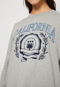 Grå sweatshirt med "California" i blå text och ett cirkulärt emblem som lyder "East Los Angeles Athletics Commons Club Sedan 1834" med lagergrenar.