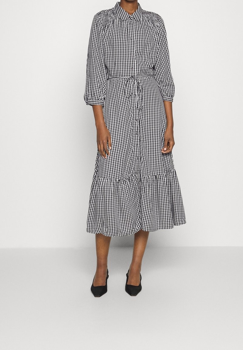 Femme portant une robe midi à boutons à carreaux vichy noir et blanc avec une ceinture nouée à la taille et des manches trois-quarts bouffantes, associée à des escarpins noirs.