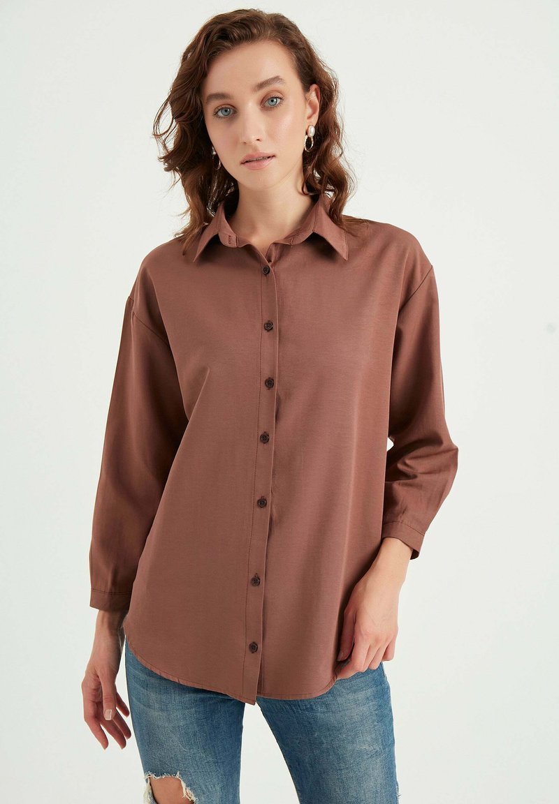 Camicia marrone con bottoni e maniche lunghe, realizzata in tessuto liscio. Presenta un colletto e un orlo arrotondato, indossata con jeans blu strappati.