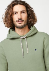 Lichtgroene hoodie van zacht materiaal, met een voorzak in kangoeroestijl, gemusterde koordjes en een klein geborduurd boombadge.
