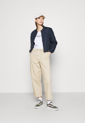 Veste bomber matelassée navy, T-shirt blanc avec logo, pantalon beigne avec cordon, baskets vertes avec accents blancs.