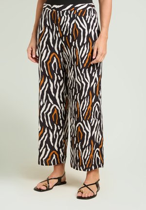 Pantaloni a gamba larga con stampa astratta animalier nera, bianca e ruggine, indossati con sandali bassi neri con cinturini, su sfondo chiaro.
