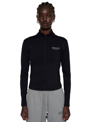Czarna kurtka na zamek z logo "HALO" noszona przez stojącą osobę w szarych dresach z małym logo na lewym udzie.