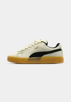 Puma Suede sportcipők krém színben, fekete oldalsó csíkkal, kerek fűzőkkel és texturált gumi talppal. Puha velúr felsőrész minimális logózással.
