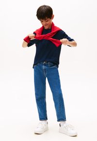Boy in blue jeans and navy shirt tying a red sweater around his shoulders, standing on a white background.